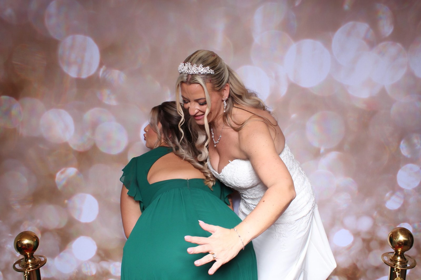 A joyful bride and bridesmaid posing candidly together on a red carpet in front of a gold sparkle photobooth backdrop at an evening wedding reception in the West Midlands
