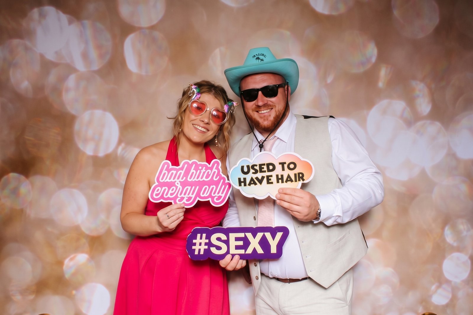 A happy wedding couple posing in front of a golden bokeh backdrop with funny custom props and novelty hats by Wards Photo Booths, Wolverhampton